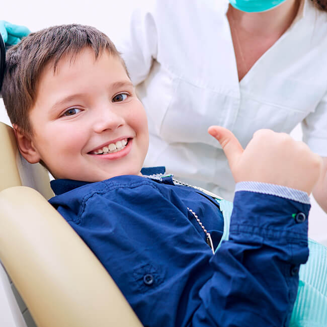 A male child sitting in a dental chair and smiling while showing thumbs up   