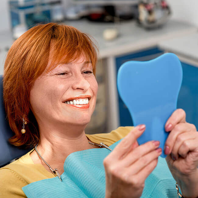 An old woman sitting in a dental chair and looking at her smile on a handheld mirror 