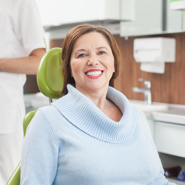 An old patient sitting in a dental chair smiling with a team member standing behind her.