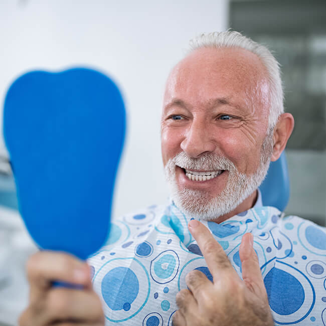 An old man sitting in a dental chair wearing a dental bib while holding a blue mirror and looking at his teeth 